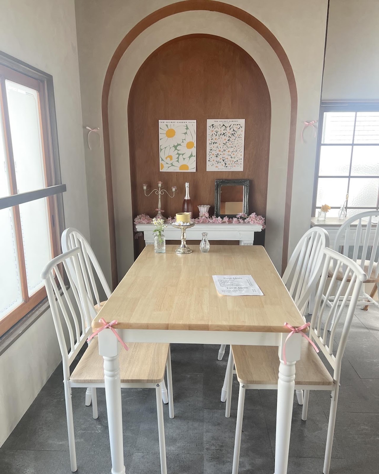 木製テーブルと白い椅子4脚が並ぶ奥の大テーブル席、正面の壁には茶色いアーチが二重に描かれ花のポスター2枚が飾られている