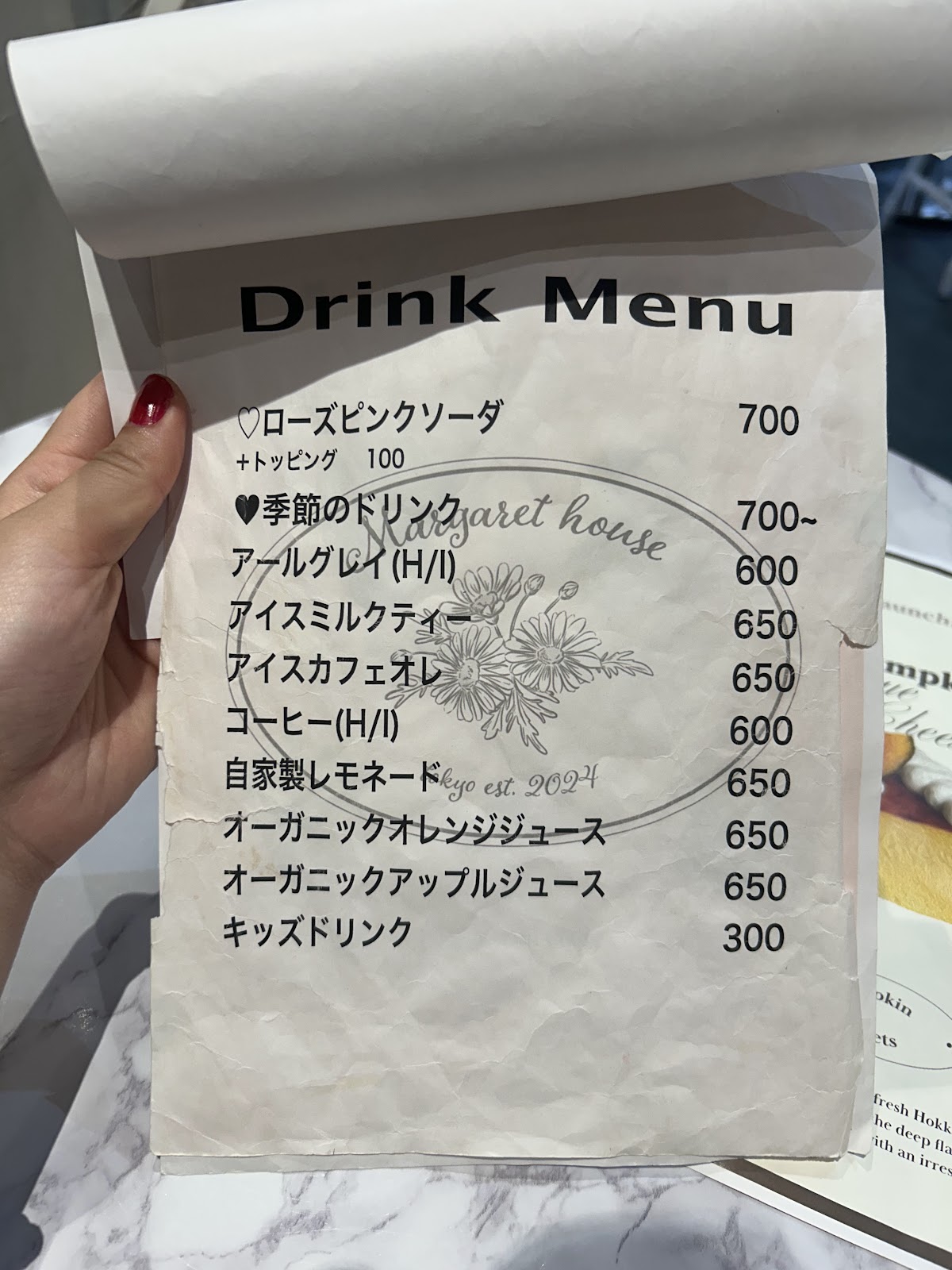 手書き風の「Drink Menu」メニュー表を手で持った写真。ローズピンクソーダ700円、アイスカフェオレ650円など価格一覧が記載されている