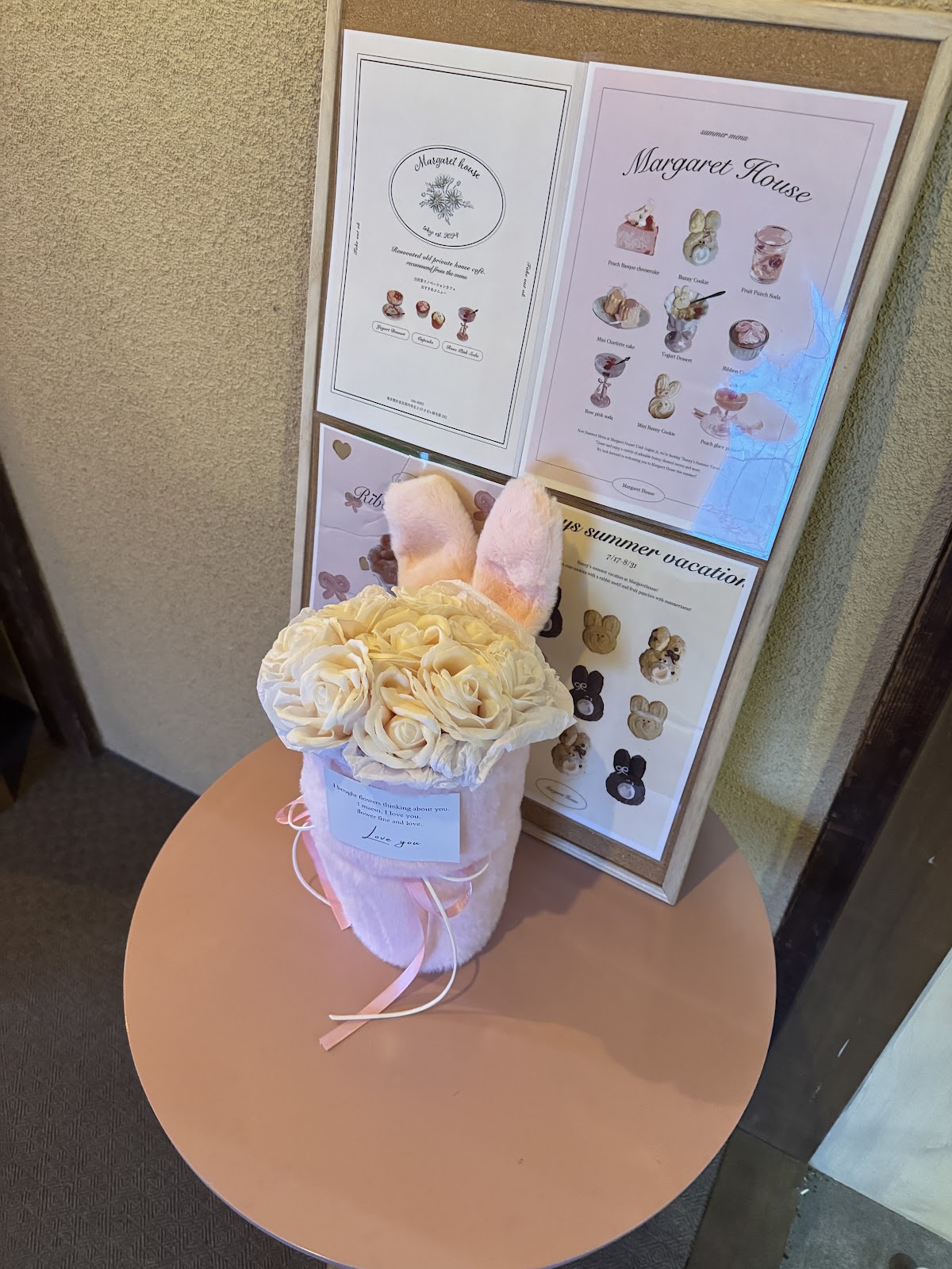 ピンクの丸テーブルの上に白いバニーぬいぐるみ付きフラワーボックスが置かれ、背後に「Margaret house」のロゴ入り案内ボードが貼られている